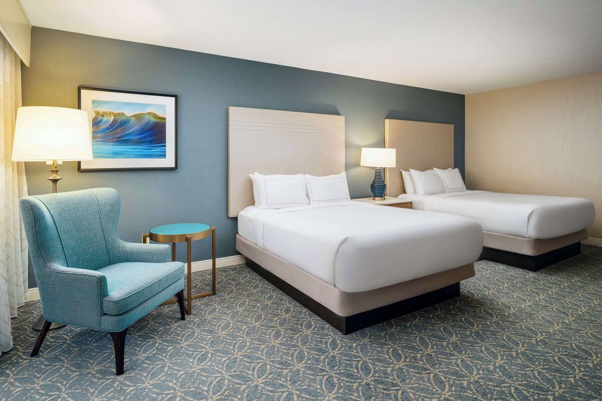 A hotel room with two beds, a blue armchair, a small round table, and a framed picture on the wall.