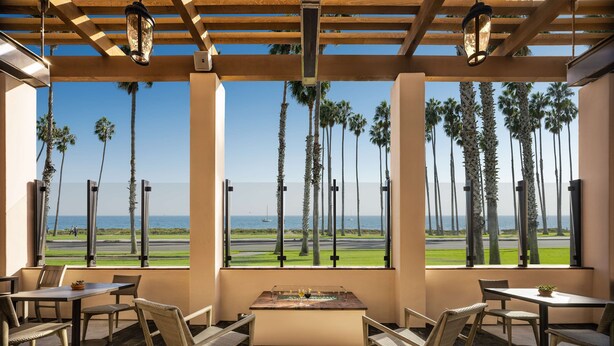 A covered outdoor seating area with tables and chairs, overlooking a runway and palm trees. - Hilton Santa Barbara Beachfront Resort (Santa Barbara)