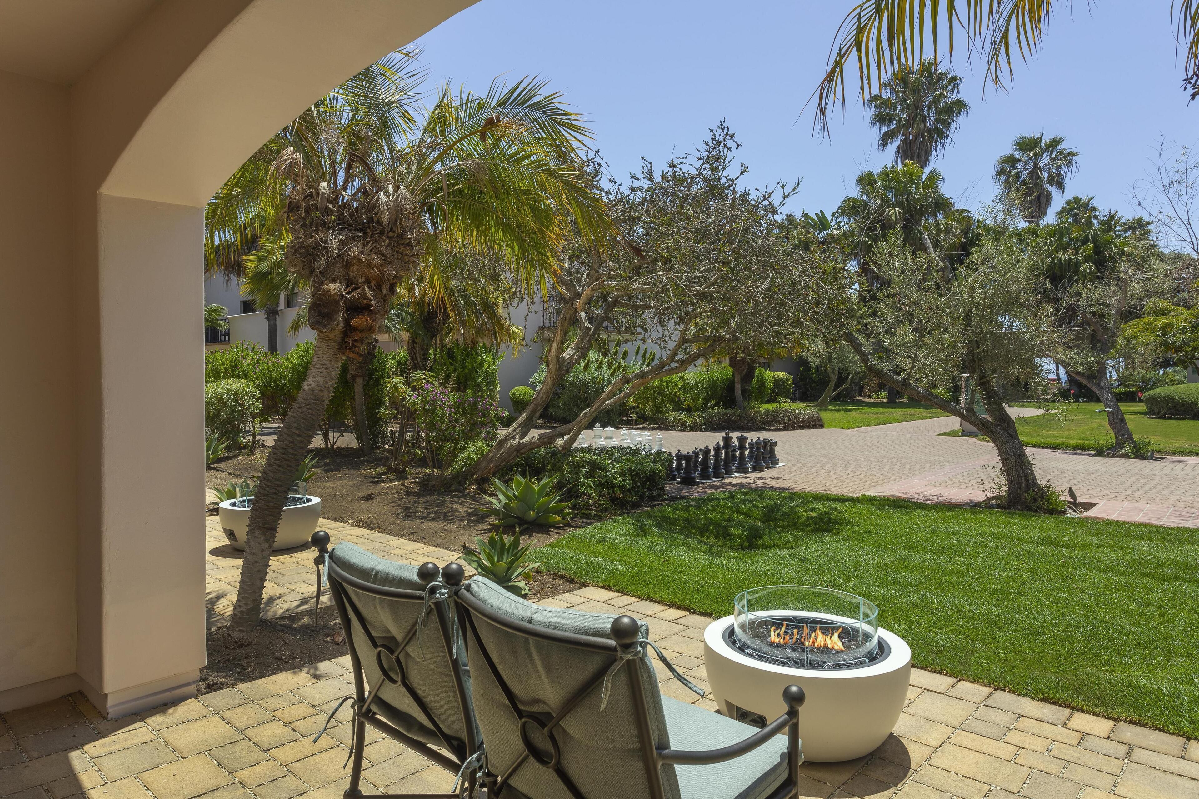 Un patio con mobiliario exterior, una chimenea y vistas a un jardín ajardinado con palmeras y un sendero.