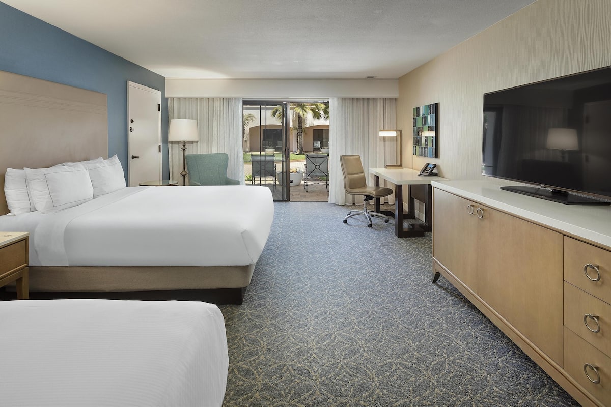 A hotel room with two beds, a desk with a chair, a TV, and a view of the outdoor area.
