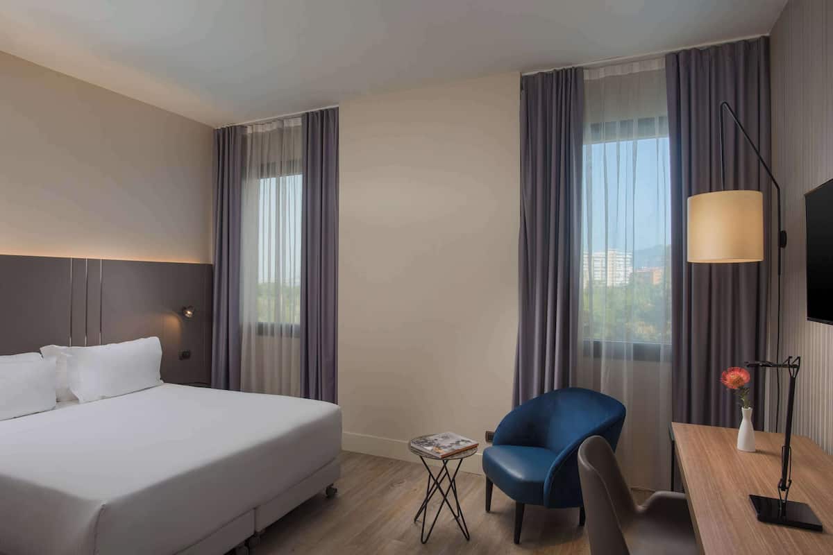 A hotel room with a bed, a desk with a lamp, a chair, and a small table with a book.