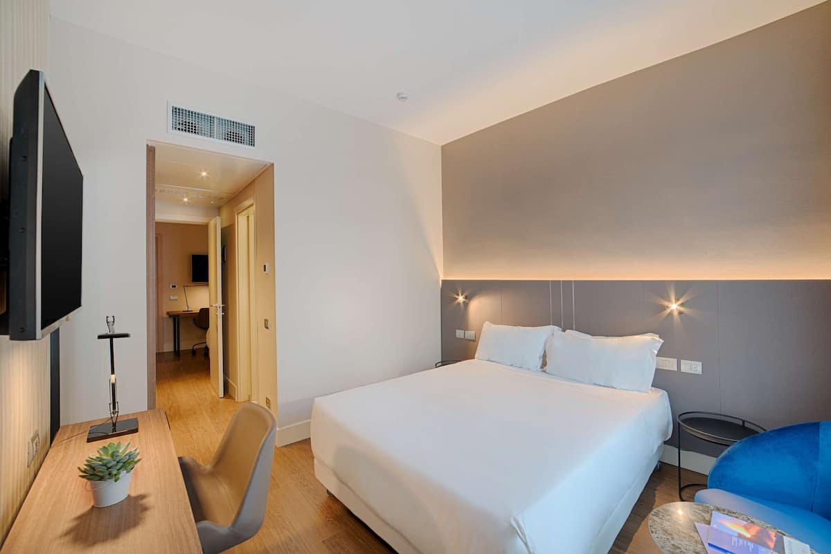 A hotel room with a bed, a desk with a plant, a TV, and a blue chair.