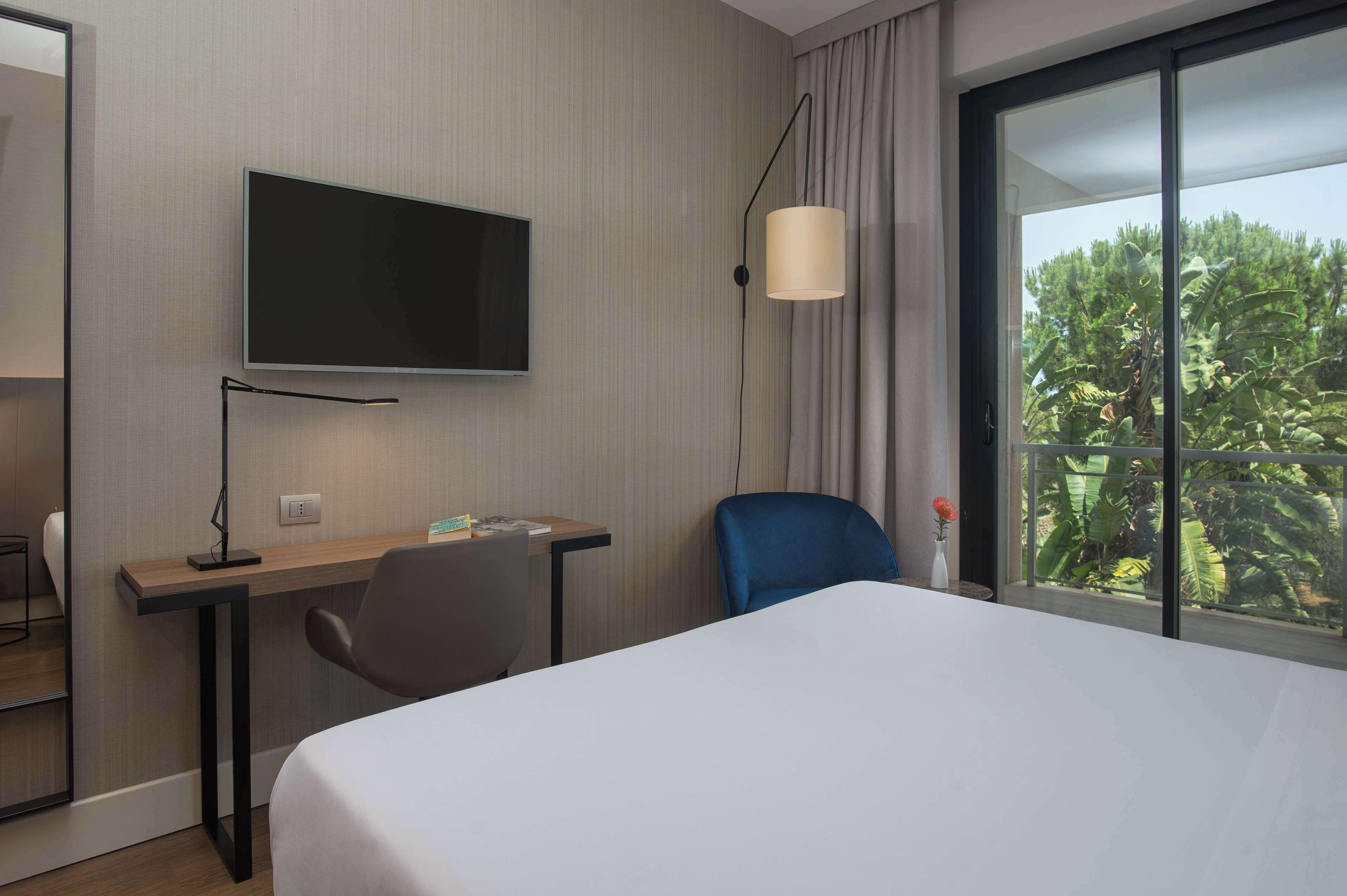 A hotel room with a bed, a desk, a chair, a TV, and a window with a view of greenery.
