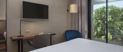 A hotel room with a bed, a desk, a chair, a TV, and a window with a view of greenery.