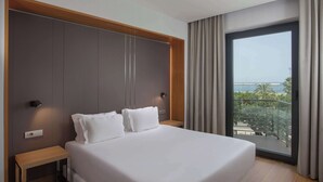 Habitación de hotel con una cama grande, una mesita de noche, una lámpara de pared y vistas al mar a través de un ventanal amplio.