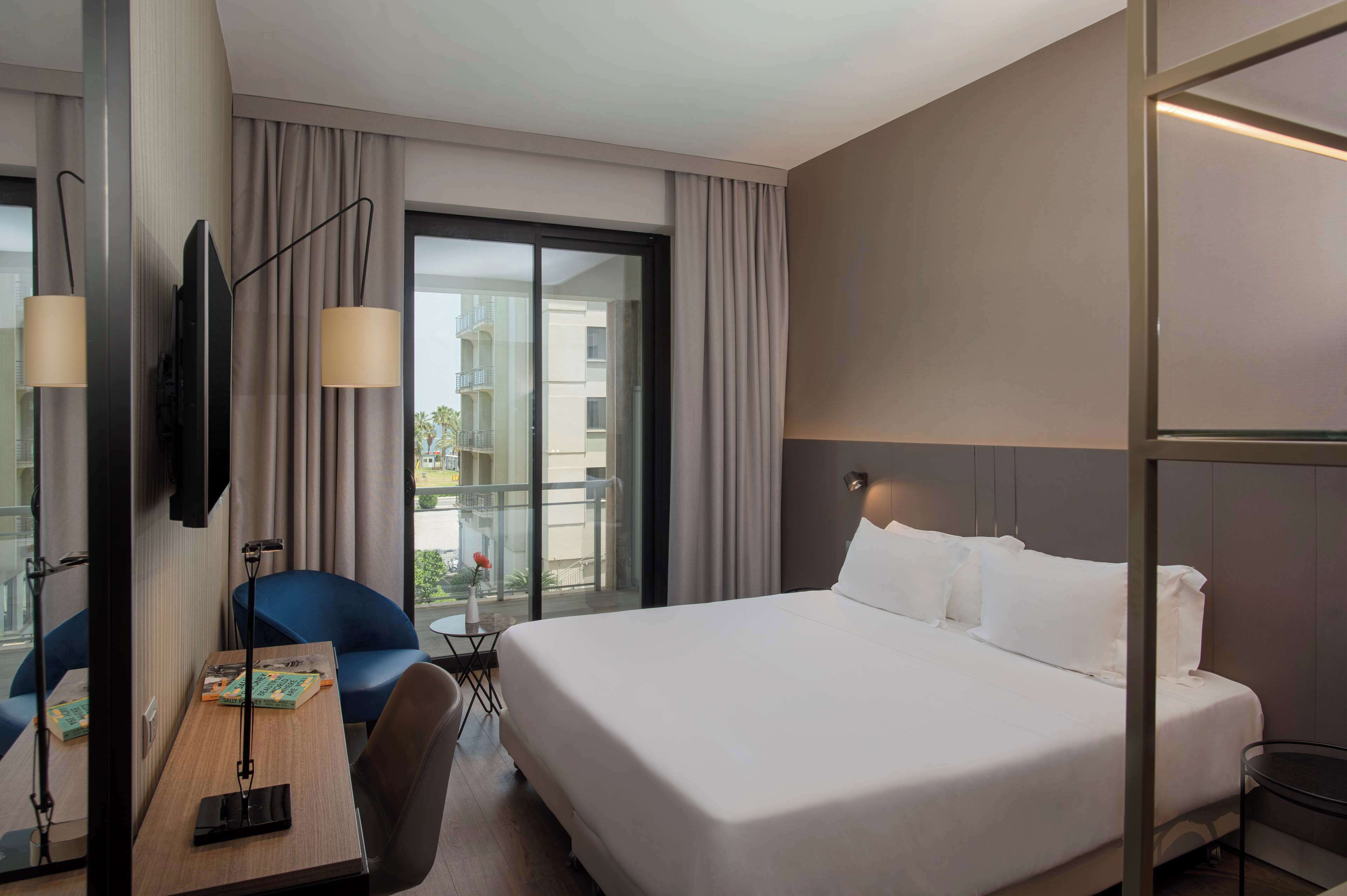 A hotel room with a large bed, a desk with a chair, a TV, and a view of the outside balcony.