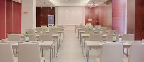 Un salón de conferencias con filas de mesas y sillas, equipado con proyectores y pantallas, y una pizarra grande.