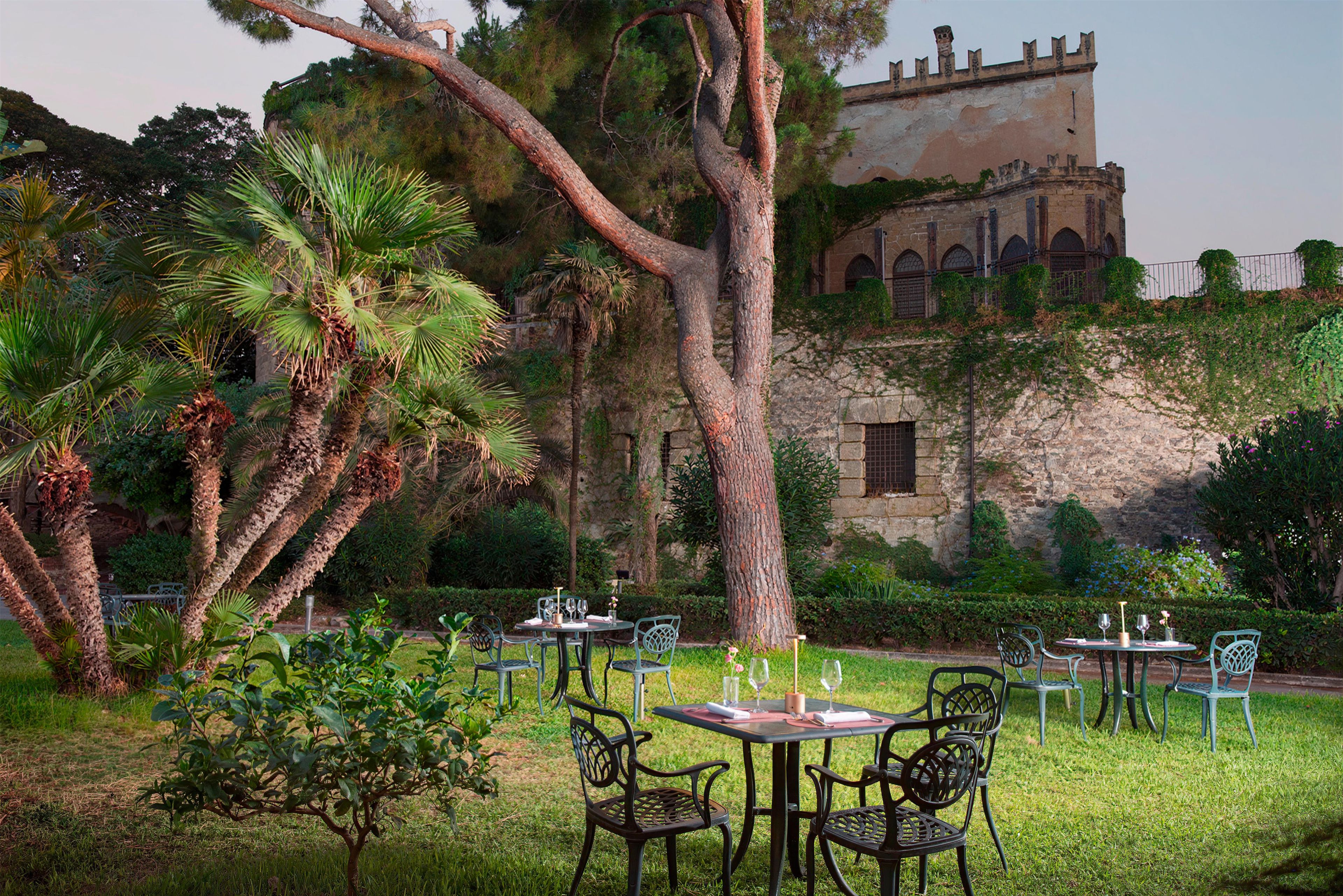 Un área de comedor al aire libre con mesas y sillas, rodeada de exuberante vegetación y un edificio histórico al fondo.
