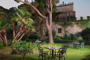 Un área de comedor al aire libre con mesas y sillas, rodeada de exuberante vegetación y un edificio histórico al fondo.