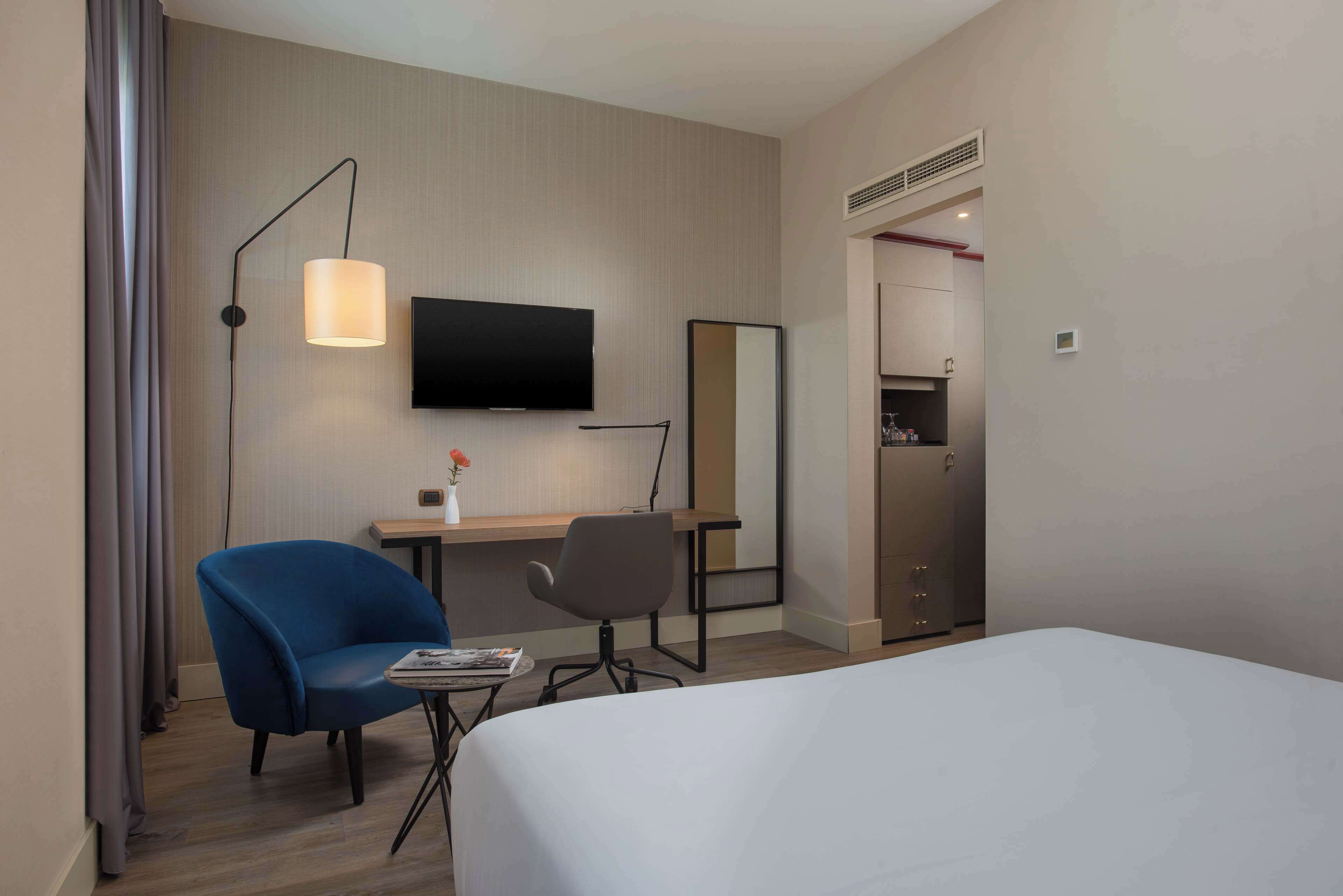 Una habitación de hotel con cama, escritorio con silla, televisión, espejo y armario.