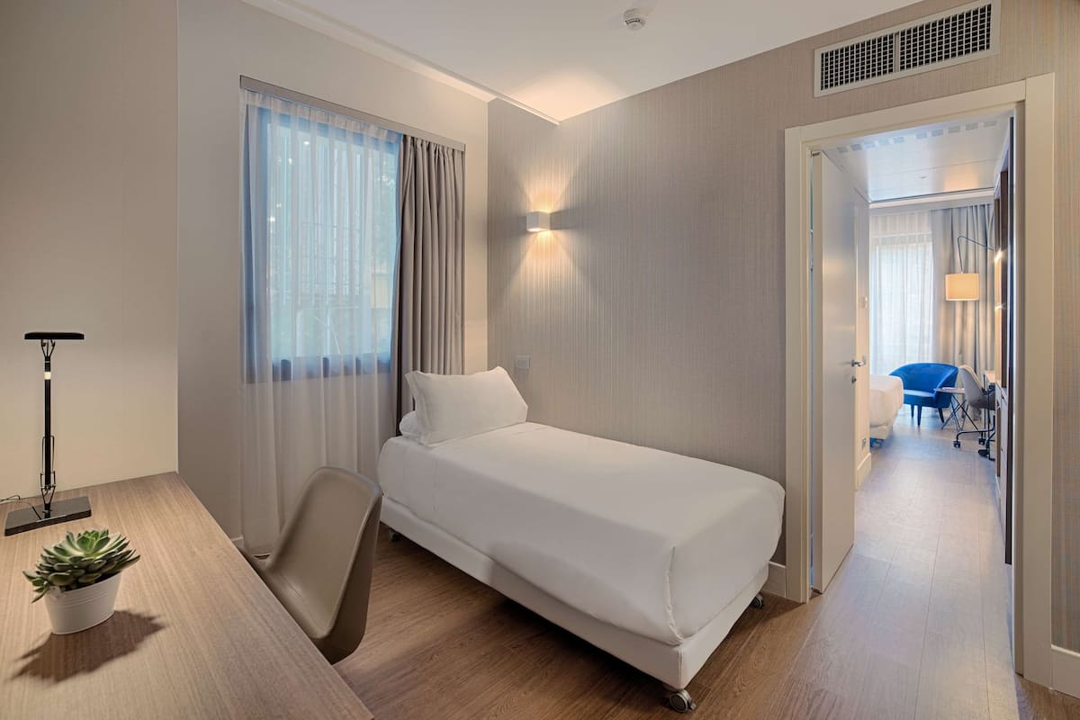 A hotel room with a single bed, a desk with a lamp, a chair, and a potted plant.