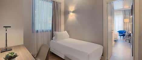 A hotel room with a single bed, a desk with a lamp, a chair, and a potted plant.
