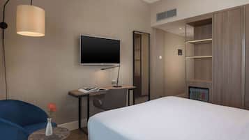A hotel room with a bed, a desk with a flat-screen TV, a chair, a small table with a vase, and a wardrobe.