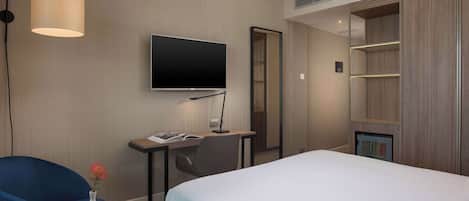 A hotel room with a bed, a desk with a flat-screen TV, a chair, a small table with a vase, and a wardrobe.