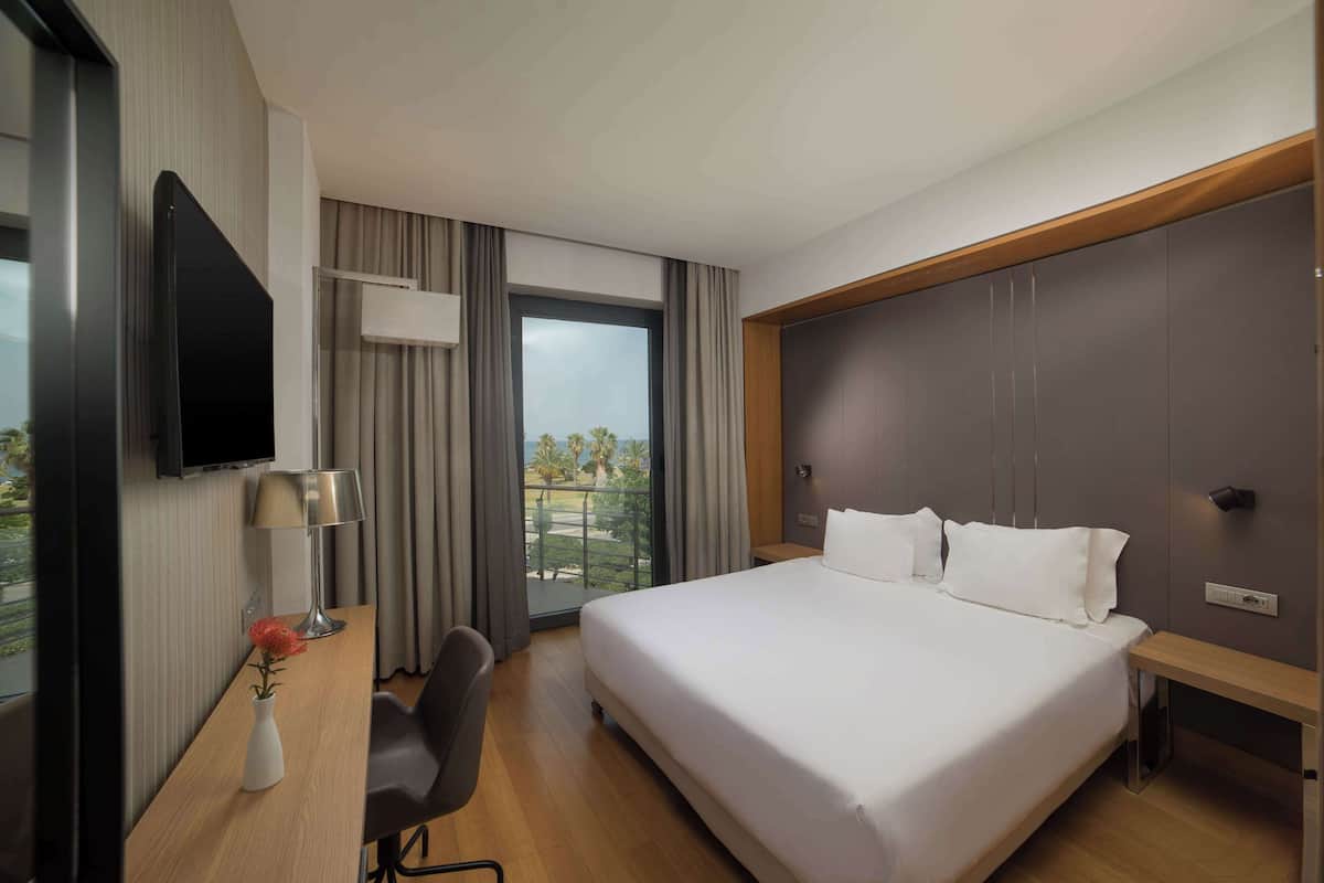 A hotel room with a large bed, a desk with a chair, a TV mounted on the wall, and a balcony with a view of palm trees.