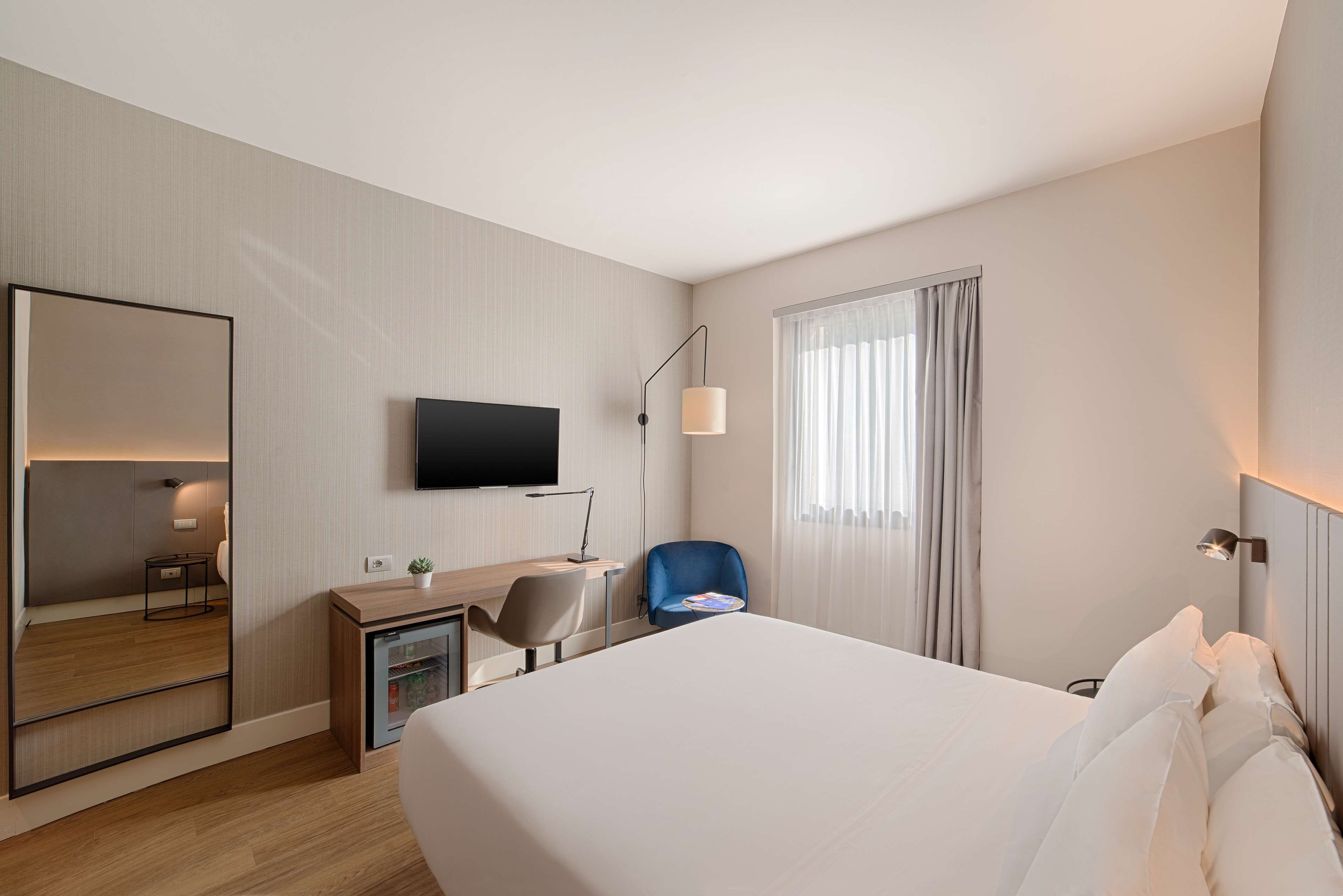 A hotel room with a large bed, a desk with a chair, a television mounted on the wall, and a window with curtains.