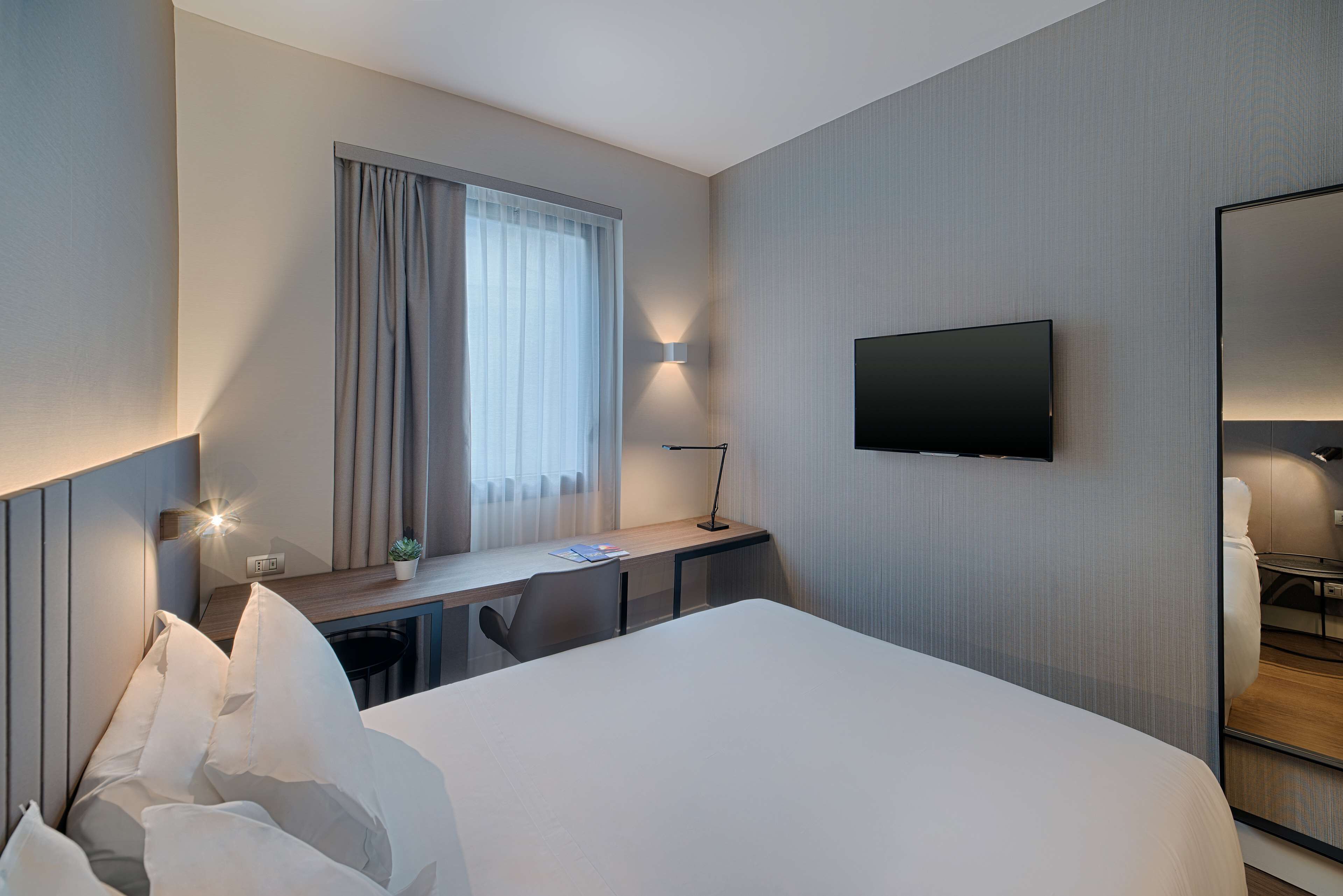 A hotel room with a bed, a desk, a chair, a TV, and a window with curtains.