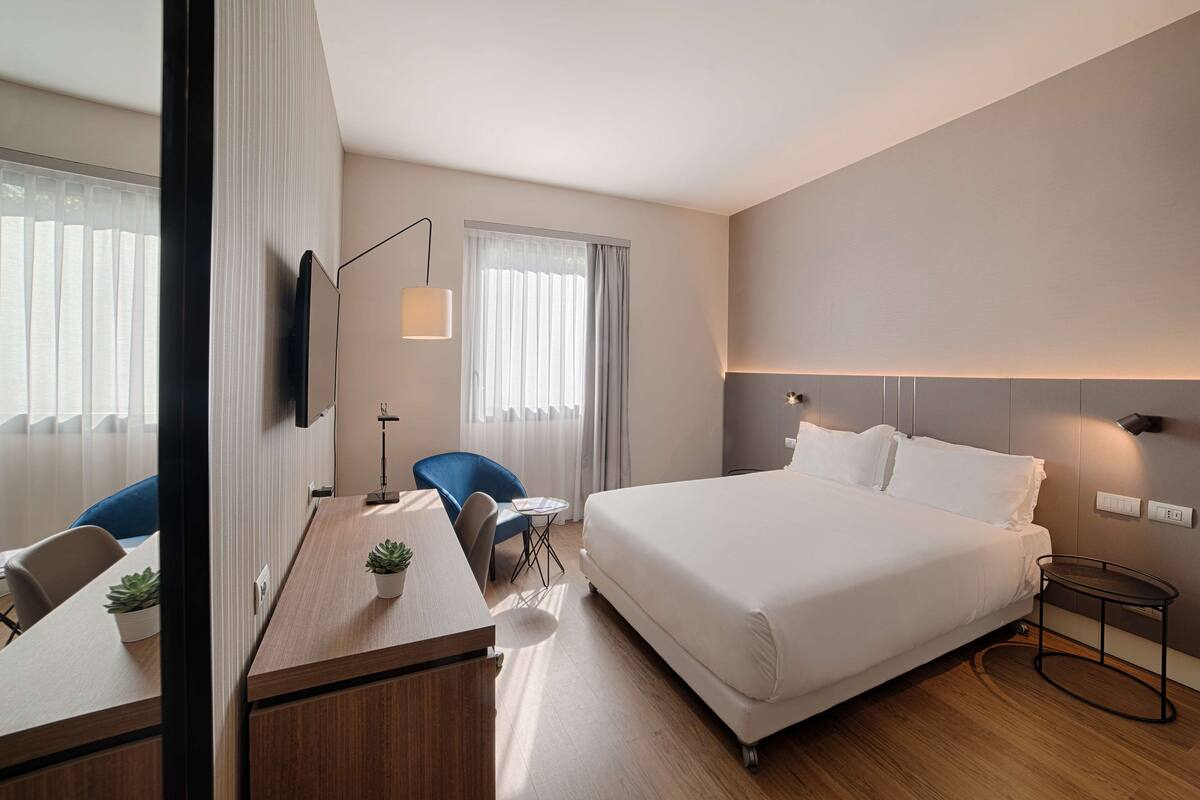 A hotel room with a large bed, a desk with a chair, a TV, and a small table with a plant.