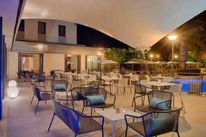 Un patio exterior con varias mesas y sillas, una piscina y un edificio moderno al fondo.