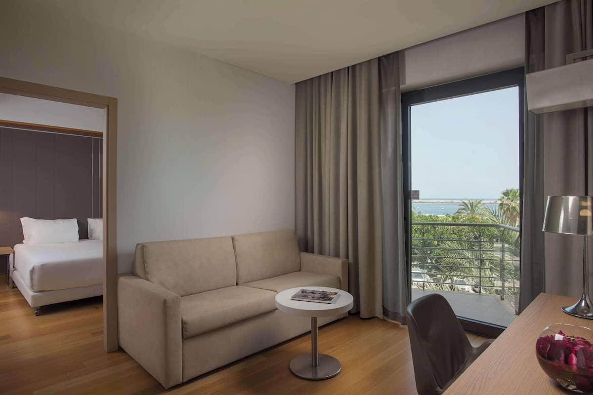 A hotel room with a bed, a sofa, a small round table, a chair, and a view of the sea through large windows.