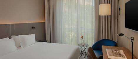 A hotel room with a large bed, a desk with a lamp, a chair, a small table with a vase, and a TV on the wall.
