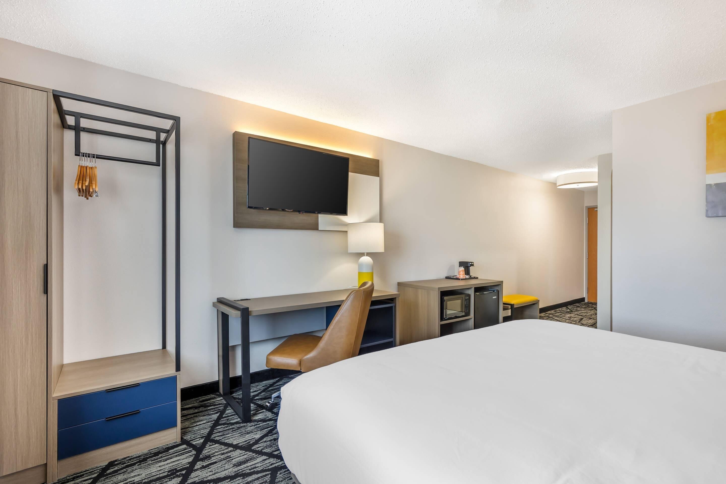A hotel room with a bed, a desk with a chair, a TV mounted on the wall, a wardrobe, and a small table with a lamp.