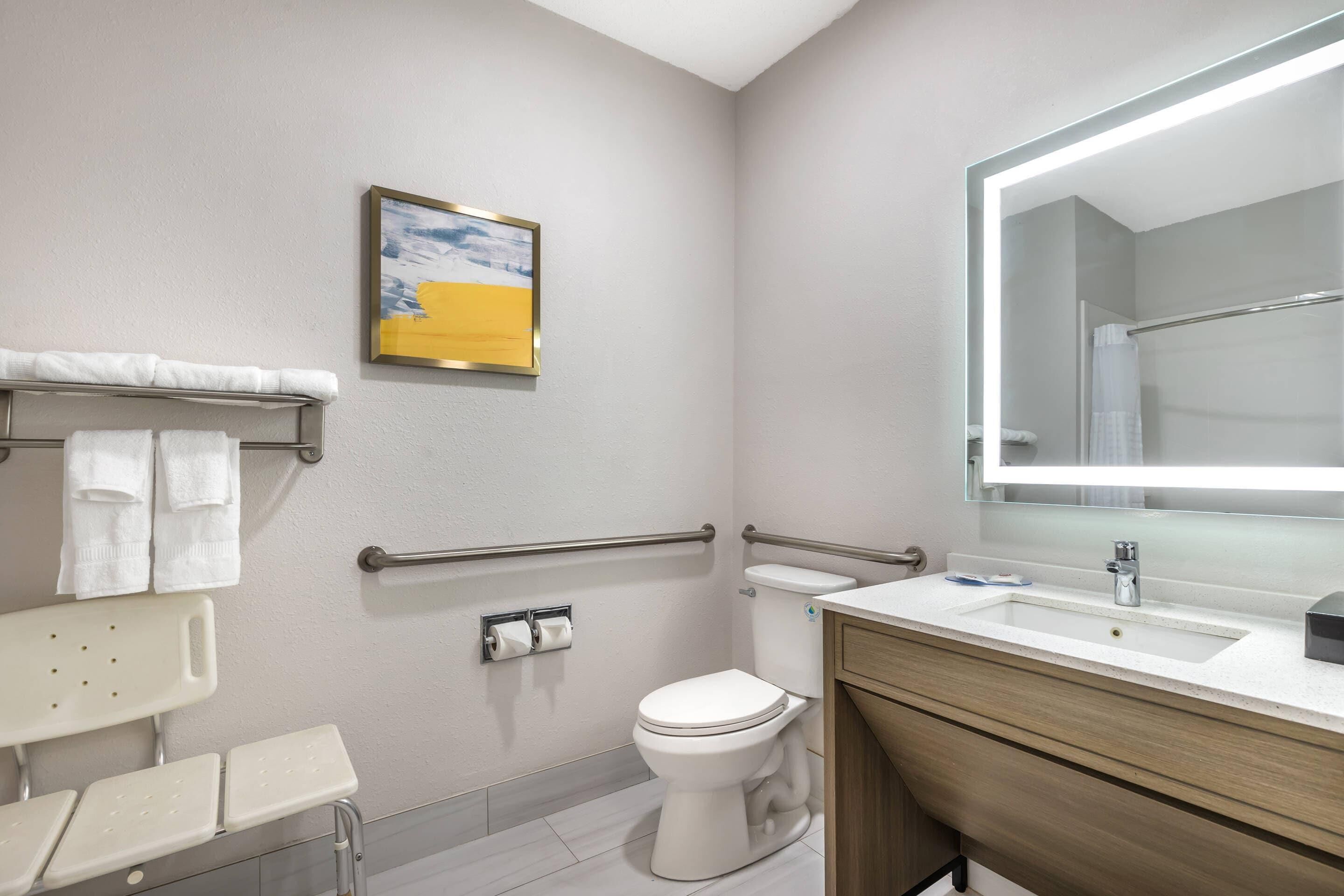 A modern bathroom with a white toilet, a sink with a mirror, a towel rack with towels, a handrail, and a painting on the wall.