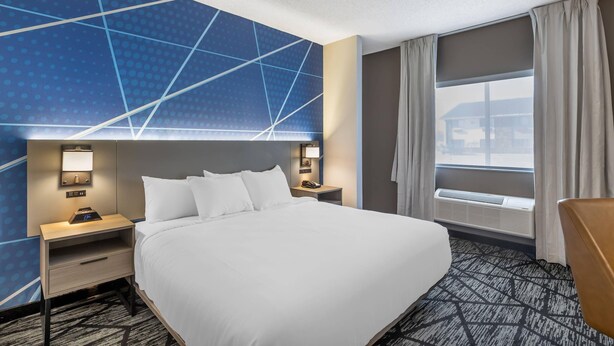 A hotel room with a large bed, a nightstand with a lamp, a desk with a phone, a window with curtains, and a view of the outside.