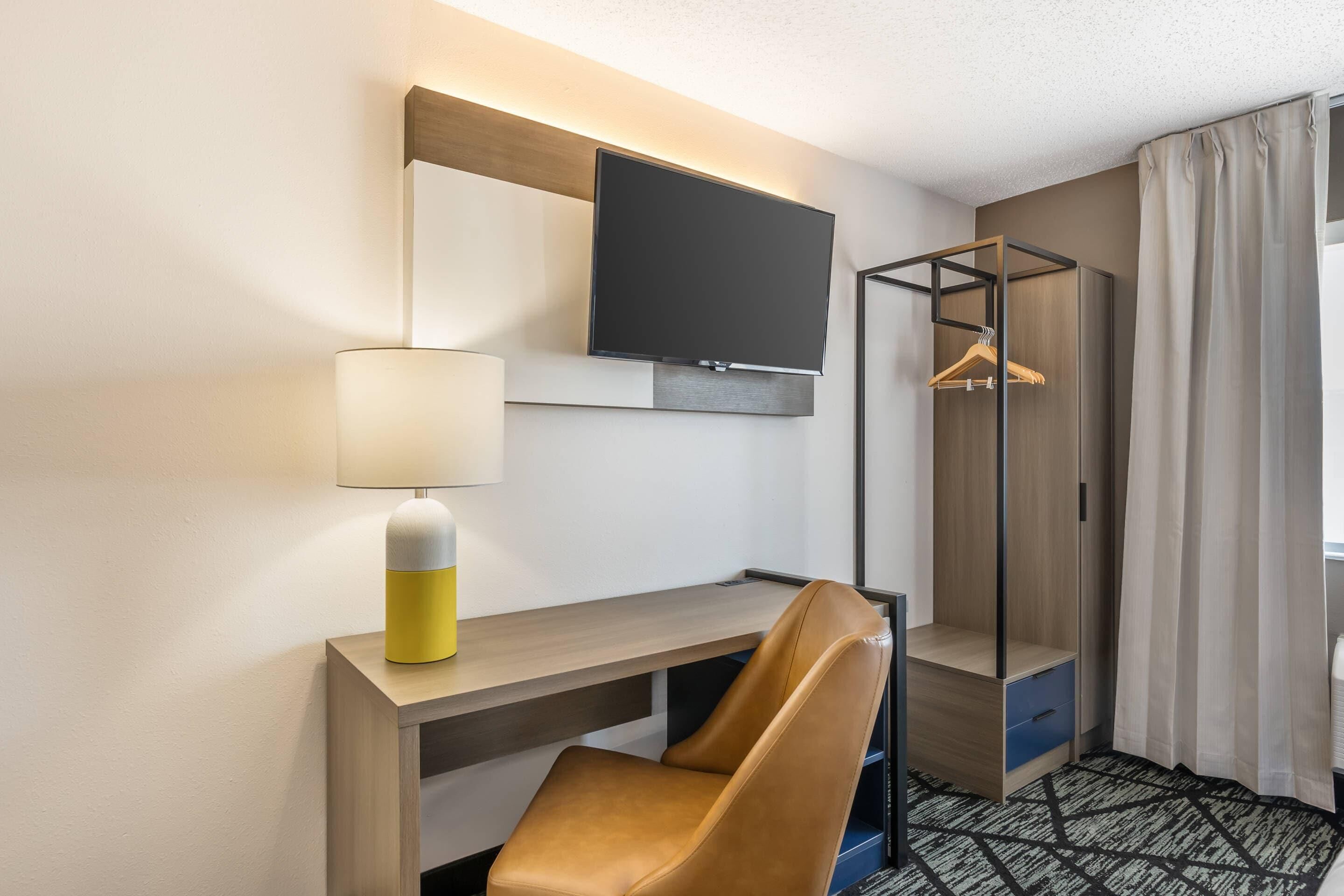 A hotel room with a desk, a television mounted on the wall, a hanging coat rack, and a bed with a patterned bedspread.