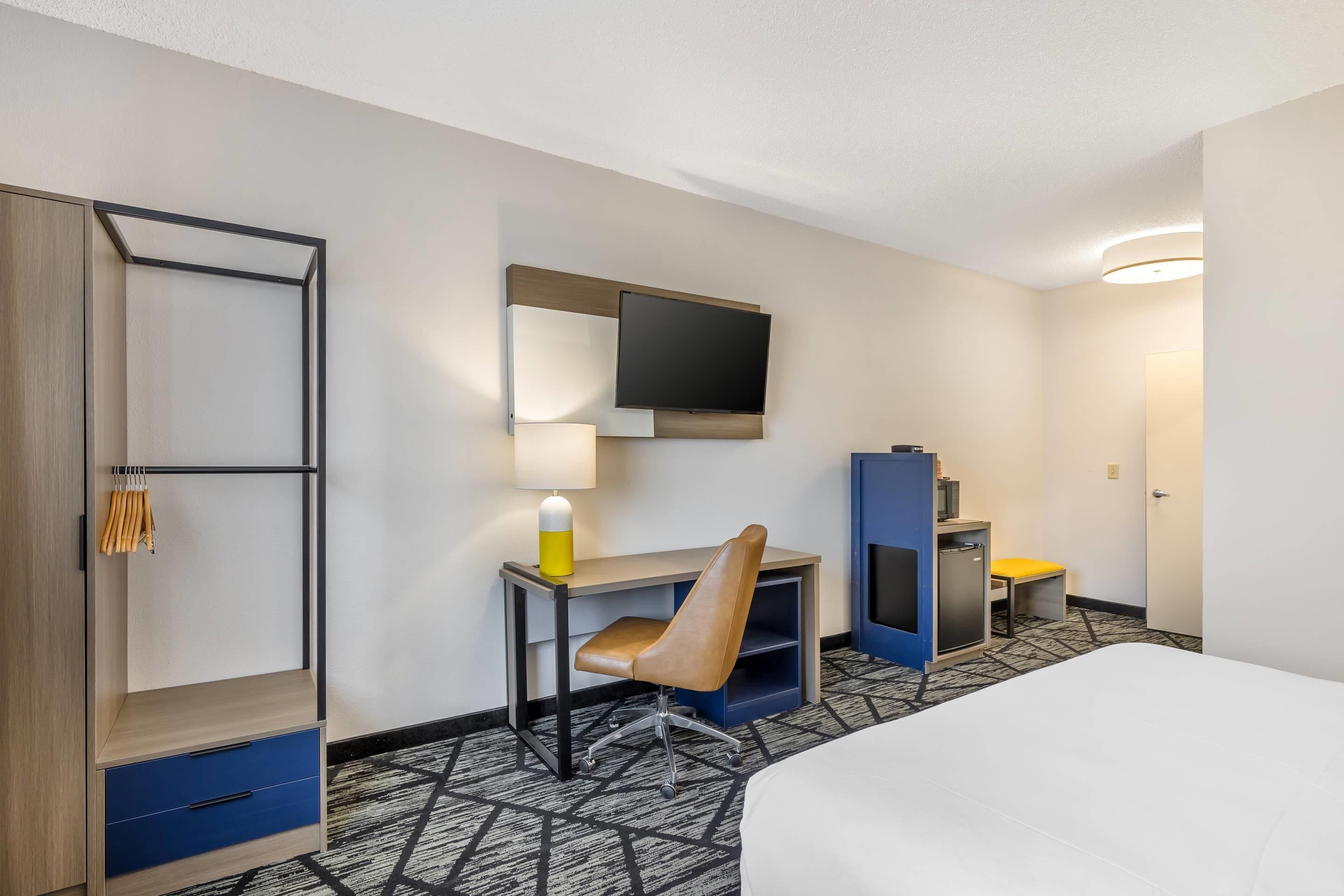 A hotel room with a bed, a desk with a chair, a TV mounted on the wall, a wardrobe, and a small table with a lamp.