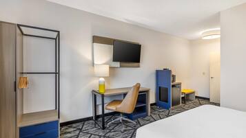 A hotel room with a bed, a desk with a chair, a TV mounted on the wall, a wardrobe, and a small table with a lamp.