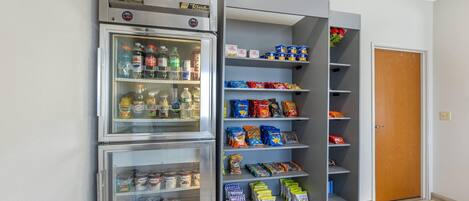 Ett litet kök med ett kylskåp fyllt med drycker och snacks, en mikrovågsugn och ett litet förråd med förpackade matvaror.