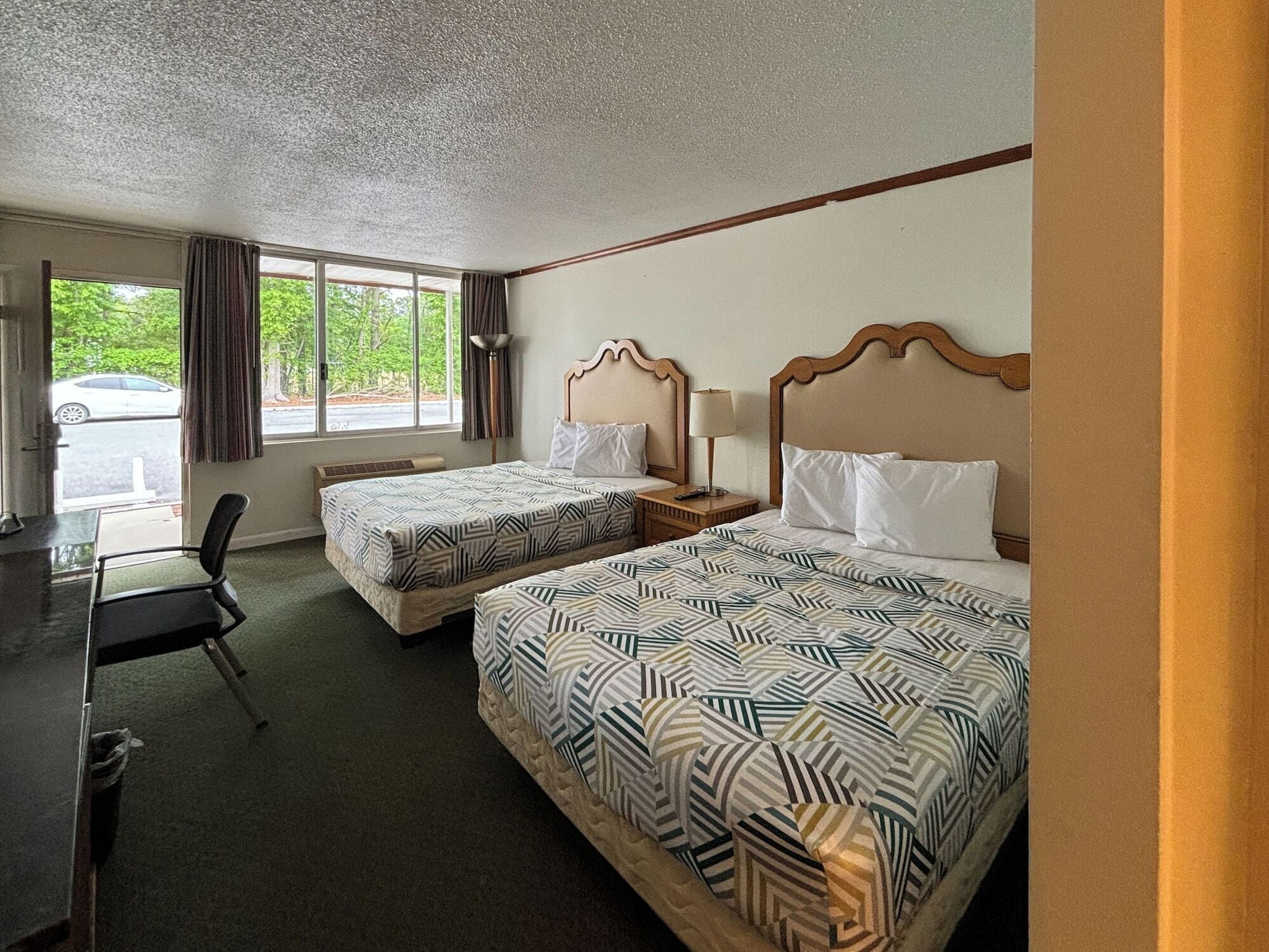 A hotel room with two beds, a desk, and a chair. There is a window with curtains and a view of the outside.
