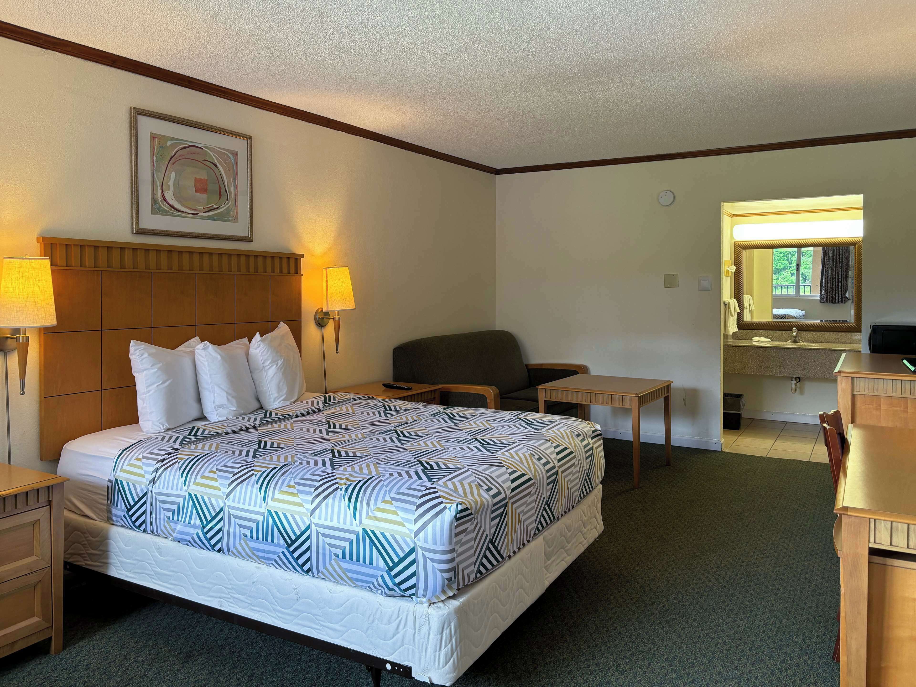 Ein Hotelzimmer mit Bett, zwei Nachttischlampen, einem Sessel, einem kleinen Tisch, einem Spiegel und einem Fenster.