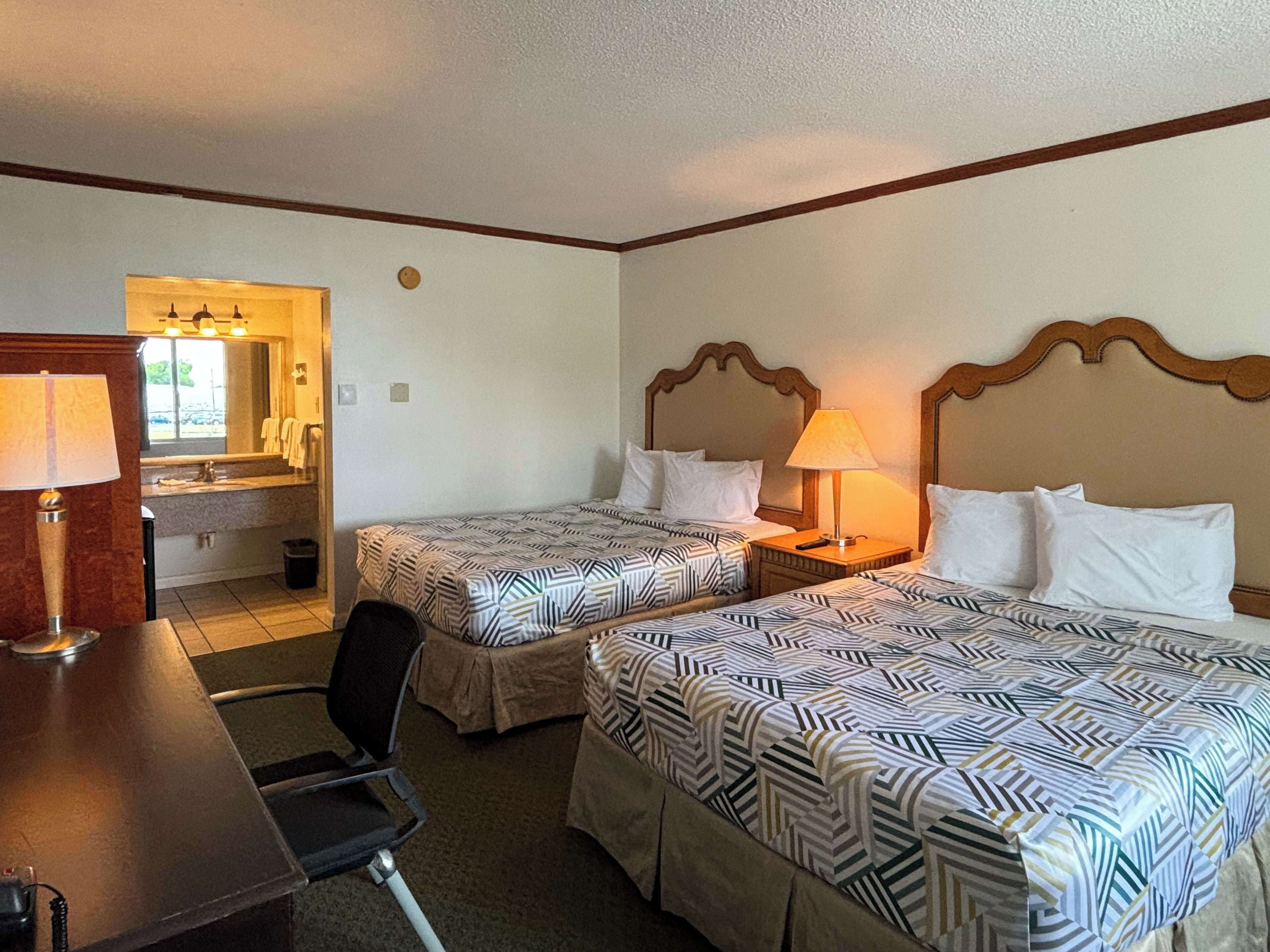 A hotel room with two beds, a desk, a chair, a lamp, and a bathroom visible through an open door.