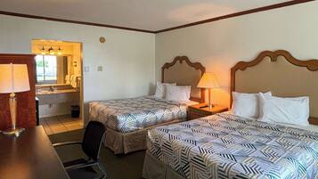 A hotel room with two beds, a desk, a chair, a lamp, and a bathroom visible through an open door.