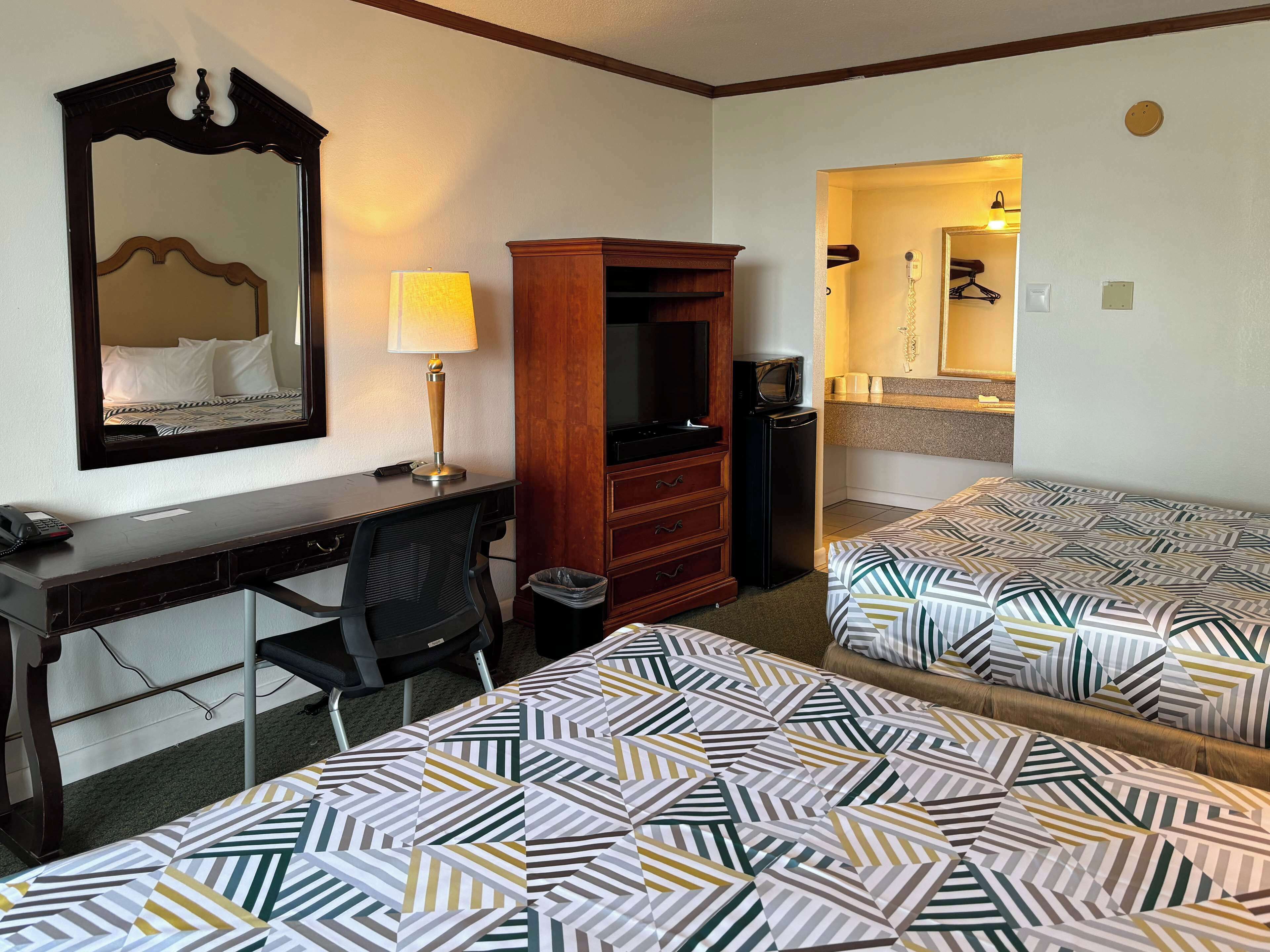Une chambre d’hôtel équipée d’un bureau, de deux lits, d’un miroir, d’une lampe, d’une armoire, d’une poubelle et d’une kitchenette.