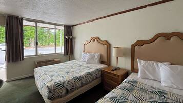 A hotel room with two beds, a window with curtains, a bedside table with a lamp, and a heater.