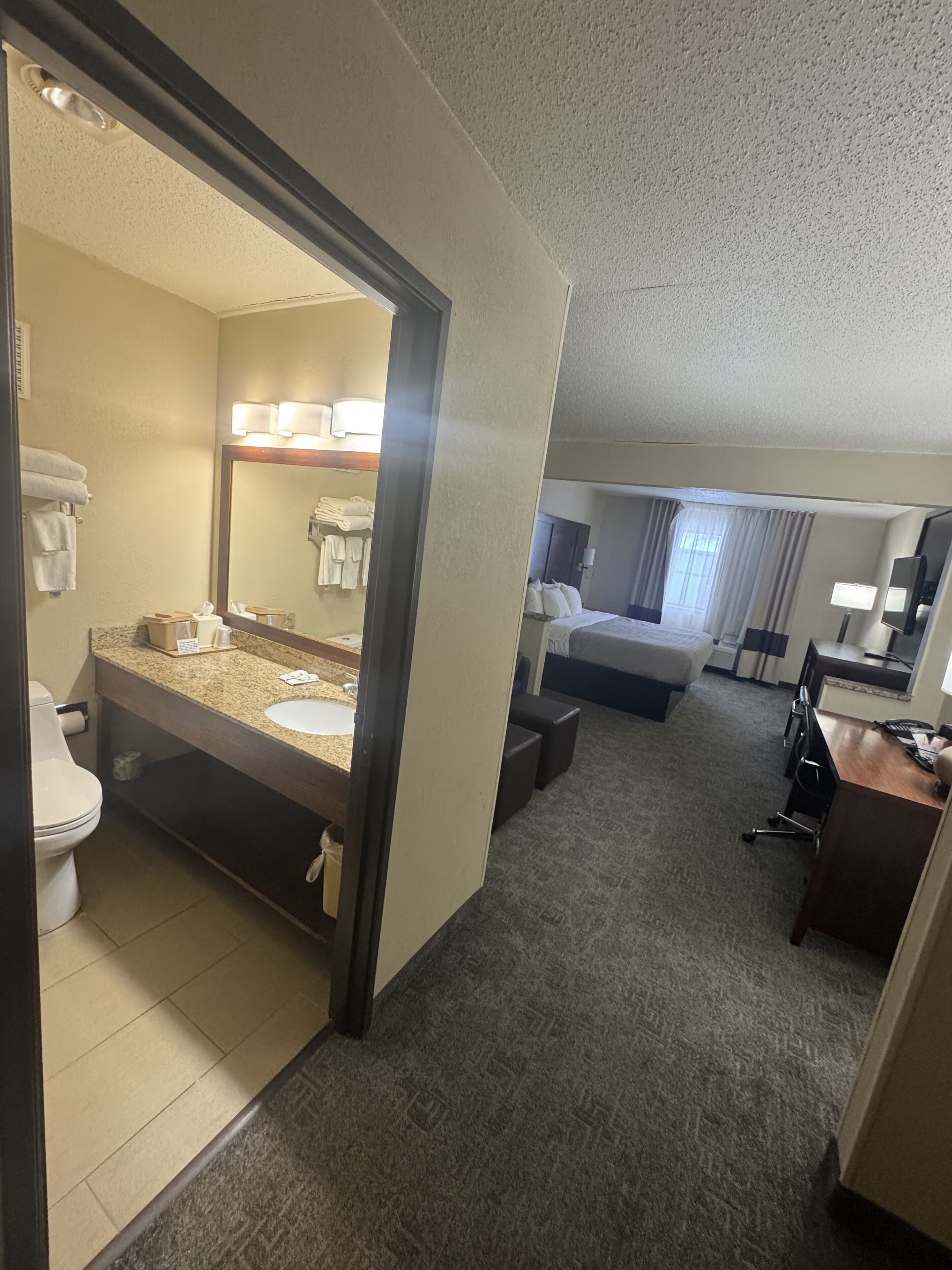 A hotel room with a bathroom, a bed, a desk, and a large mirror.