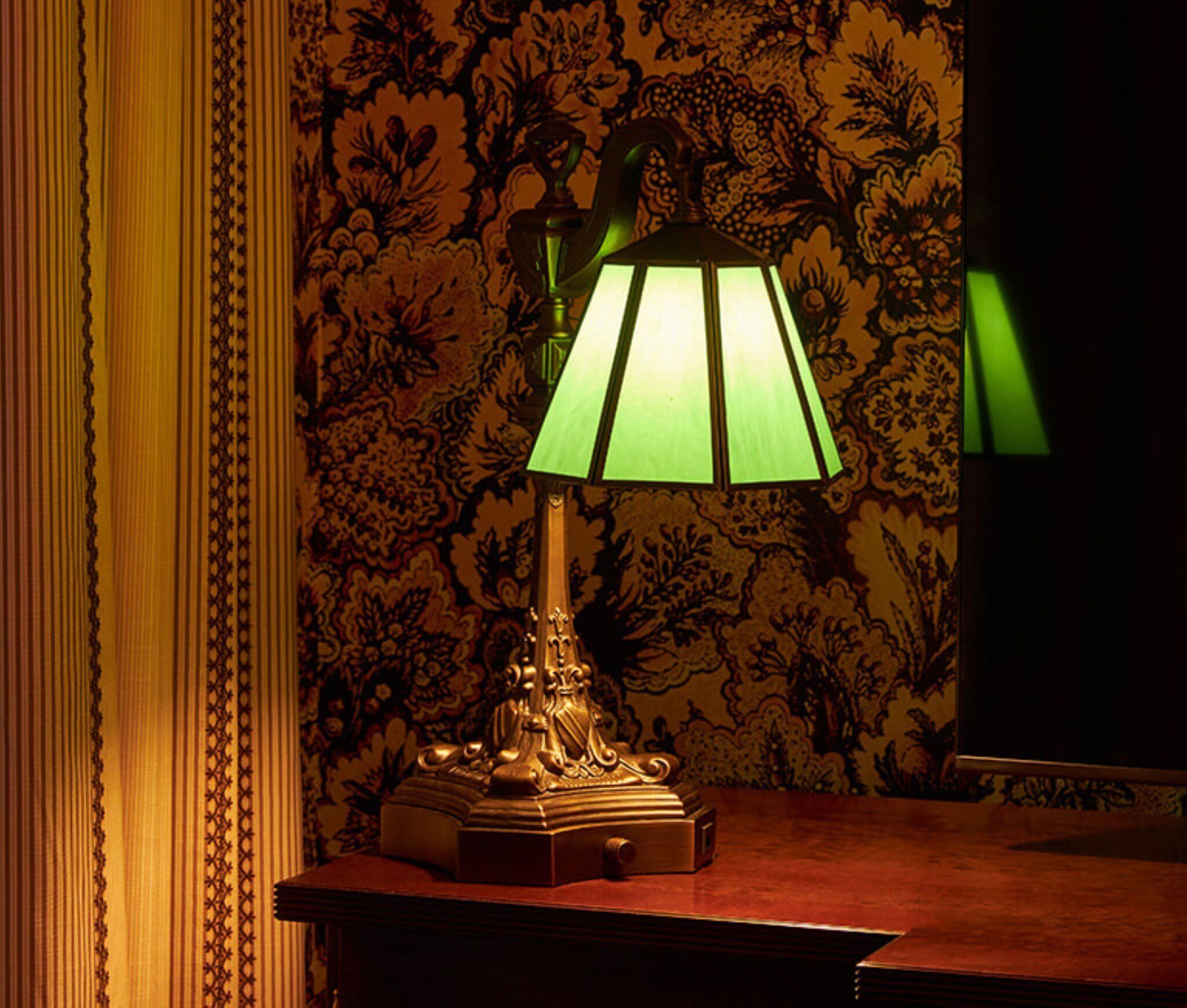 Une lampe de table avec un abat-jour vert, posée sur une table en bois, le tout sur fond d’un papier peint à motifs floraux.