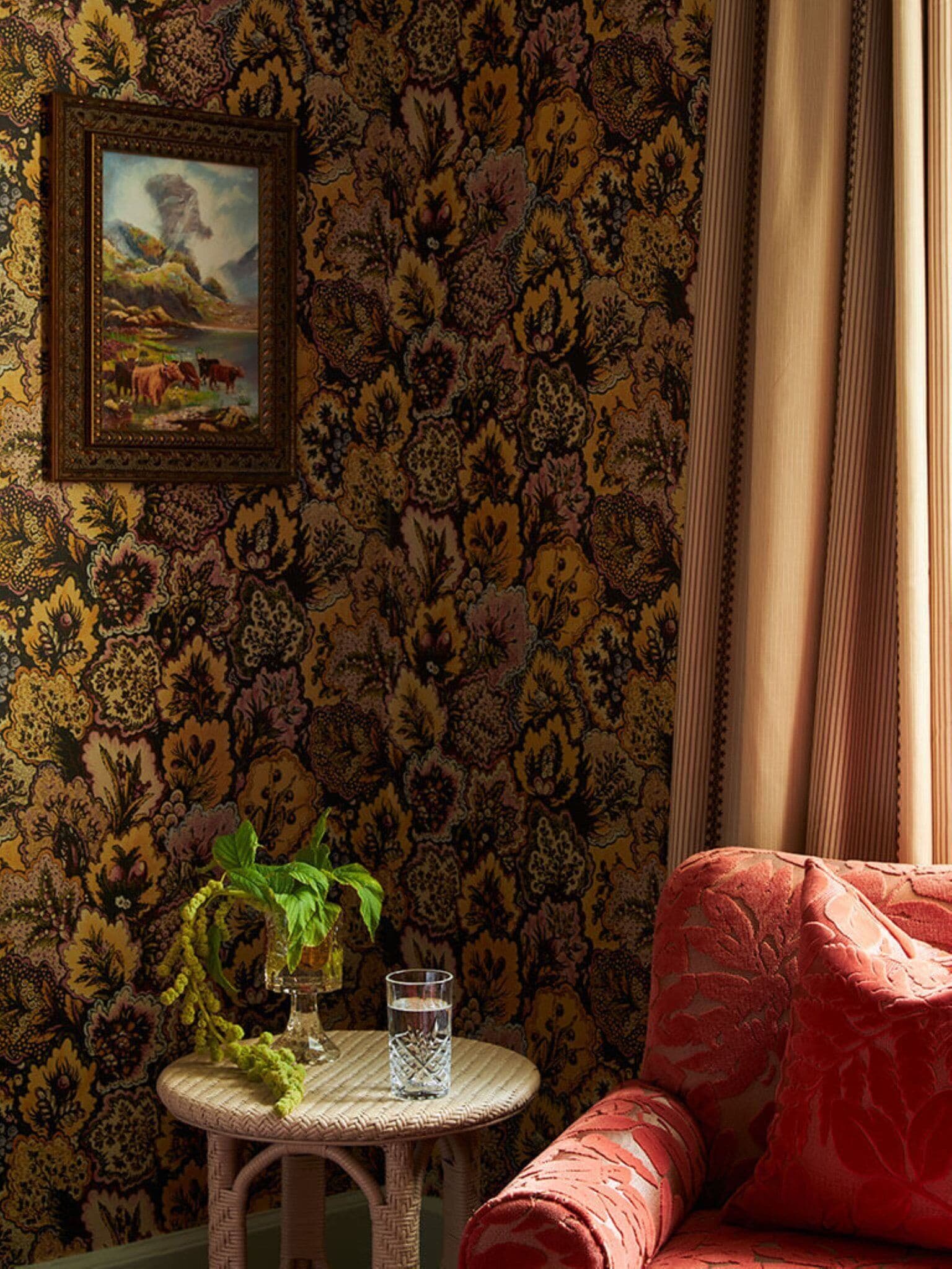 A room with patterned wallpaper, a wicker table with a glass and plant, and a red upholstered chair.