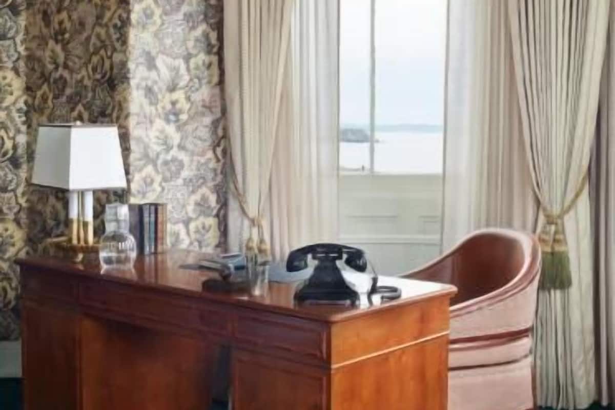 Um quarto com escrivaninha de madeira, uma cadeira, um abajur e uma janela com cortinas.