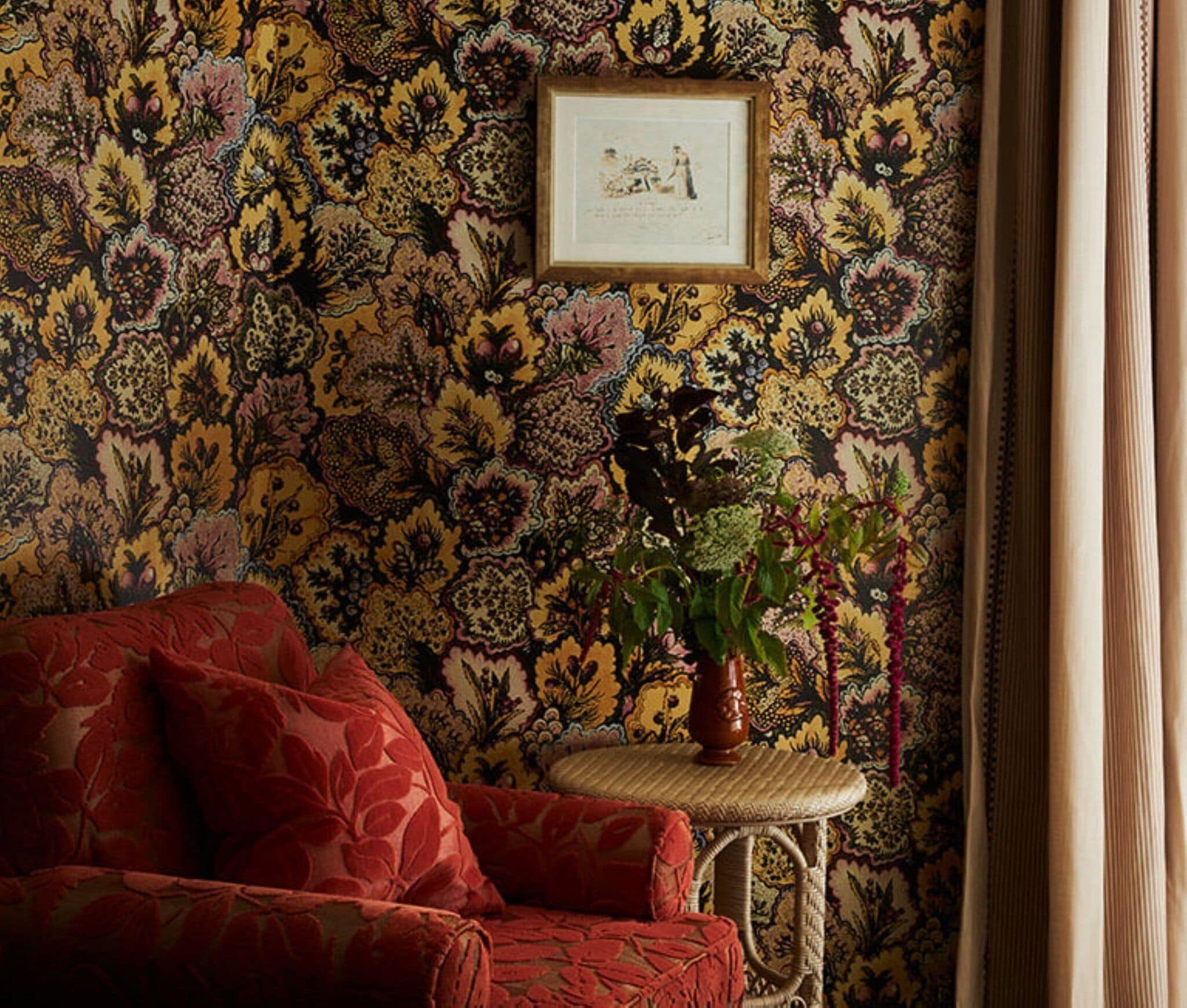 A room with a floral wallpaper, a red sofa, a small wicker table with a vase, and a framed picture on the wall.