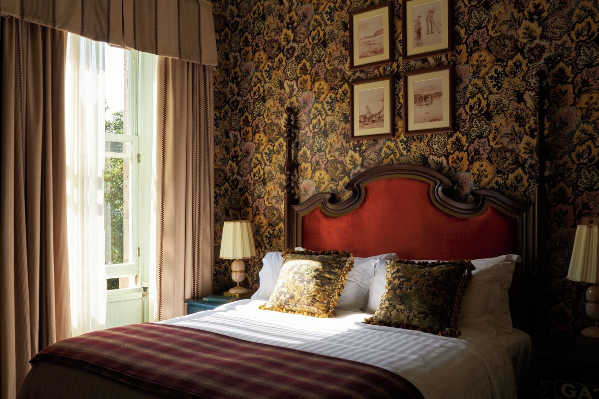 A bedroom with a large bed, ornate headboard, floral wallpaper, and framed pictures on the wall.
