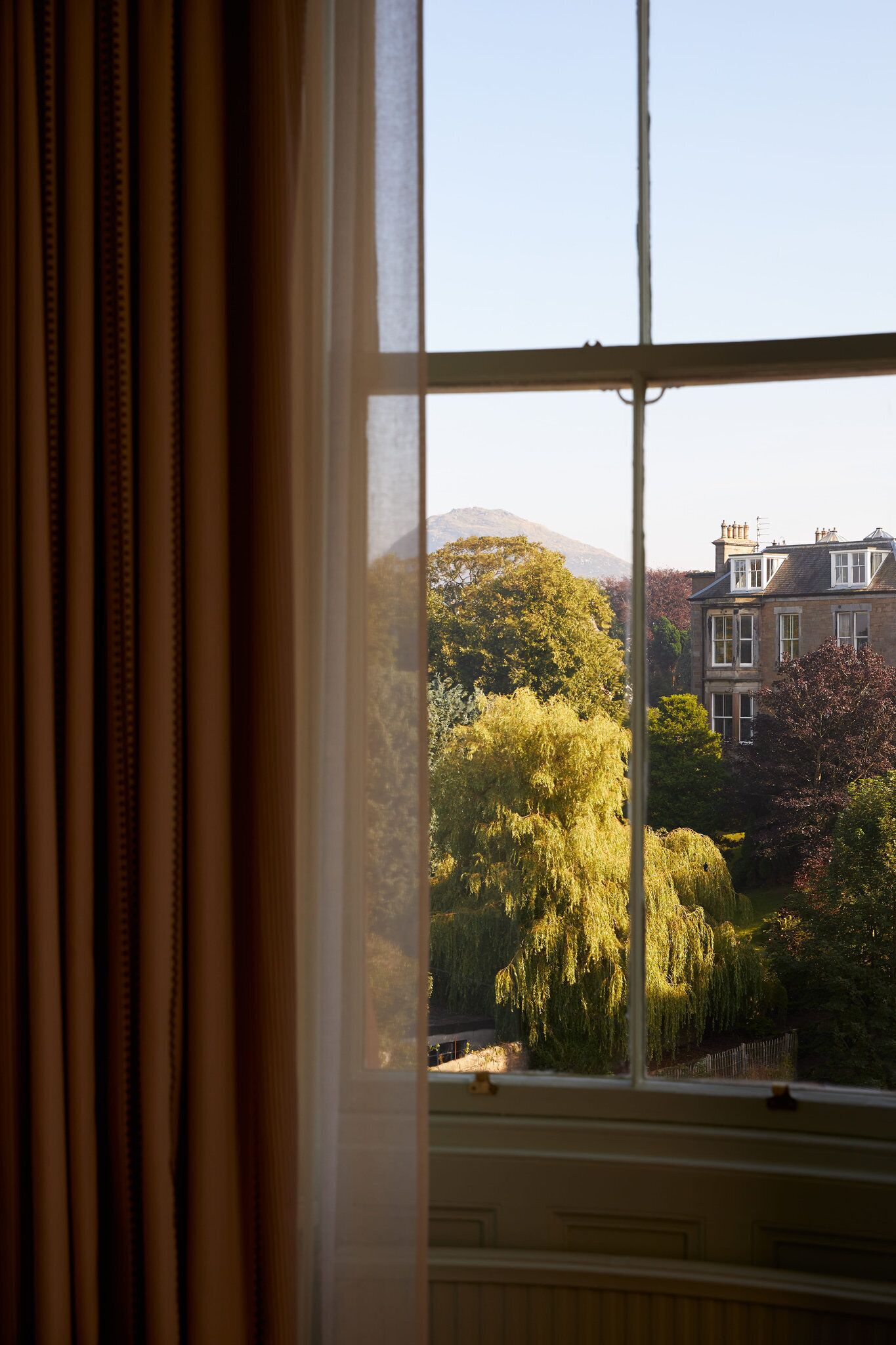 Una ventana con cortinas, vista de una casa con jardín y una montaña al fondo.