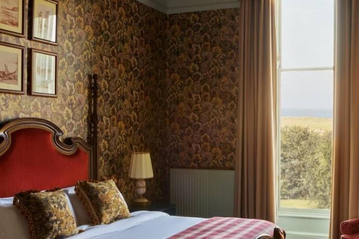 Um quarto com cama, uma cadeira vermelha, janela com vista e papel de parede estampado.