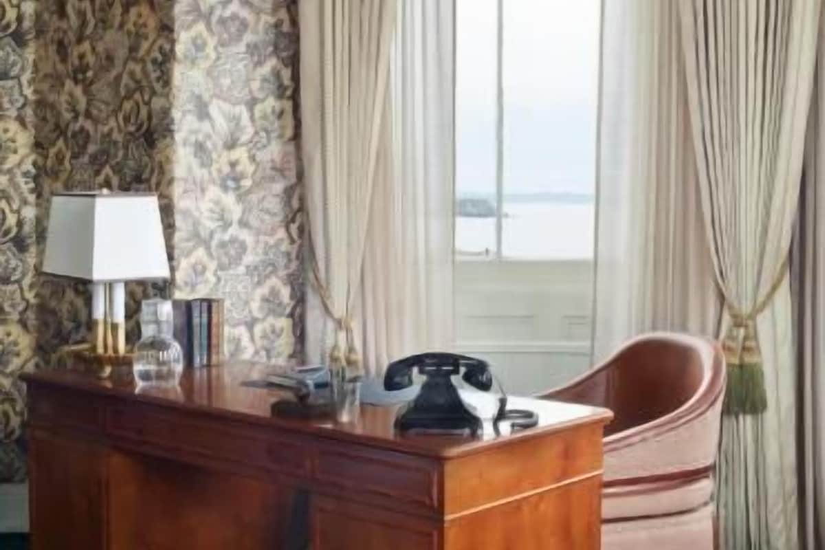 Une pièce avec un bureau en bois, une chaise, une lampe et une fenêtre avec des rideaux.