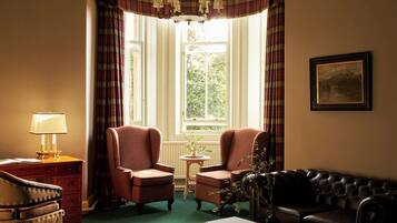 A room with a green carpet, two armchairs, a black sofa, a wooden side table with a book, a lamp, and a framed picture on the wall.