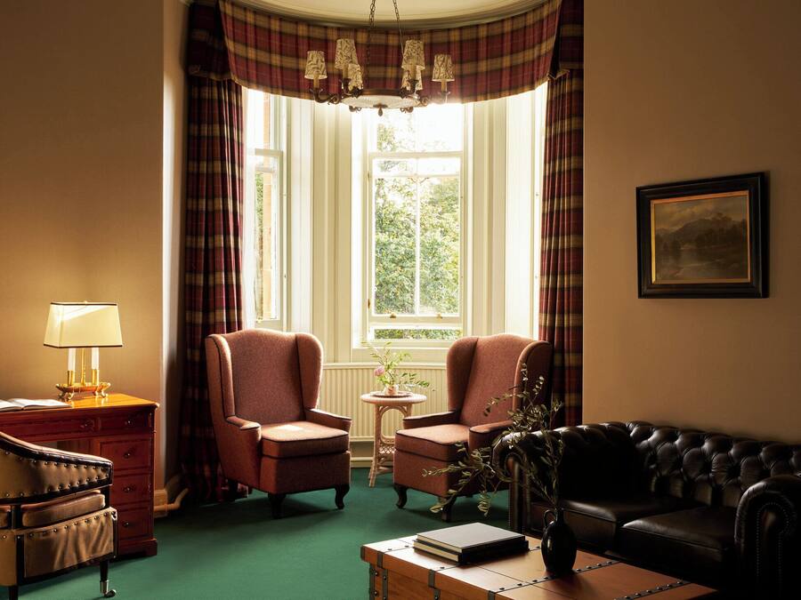 A room with a green carpet, two armchairs, a black sofa, a wooden side table with a book, a lamp, and a framed picture on the wall.
