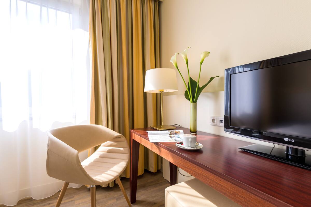 A hotel room with a wooden desk, a television, a lamp, a vase with flowers, a chair, and a window with curtains.